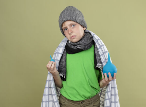 不高兴生病的小男孩戴着暖和的帽子 围着围巾 裹着毯子 拿着灌肠剂 站在轻薄的墙上 洛克感到困惑和不快温暖毯子少