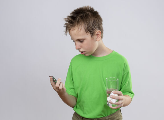 疾病生病的小男孩穿着绿色t恤感到不舒服拿着一杯水看着站在白墙上的药丸药丸不舒服感觉