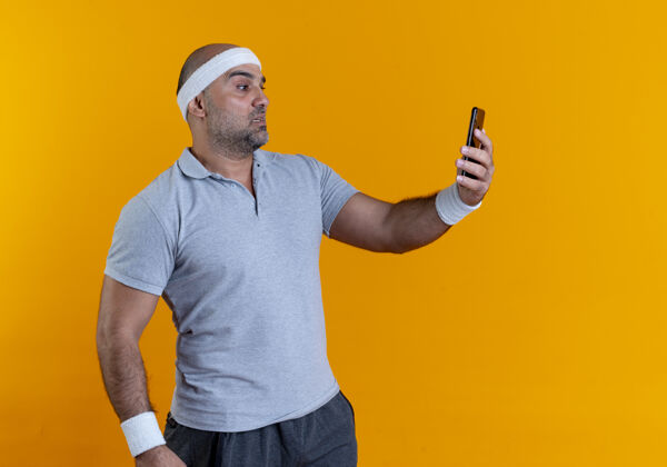 看戴着头巾的成熟运动型男人站在橙色的墙上用智能手机自拍 看起来很困惑运动运动装自拍