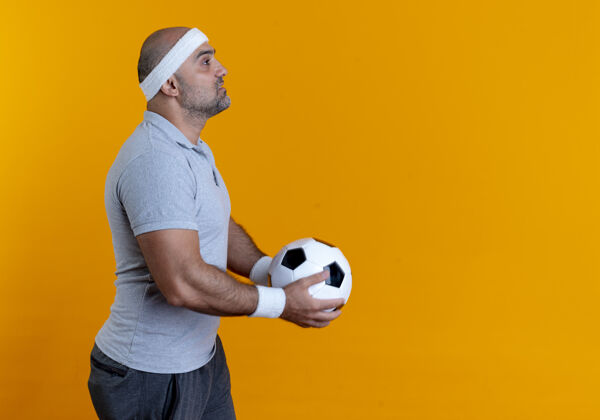 头带成熟的运动型男人戴着头巾 举着足球站在一边 严肃的脸越过橙色的墙姿势男人严肃