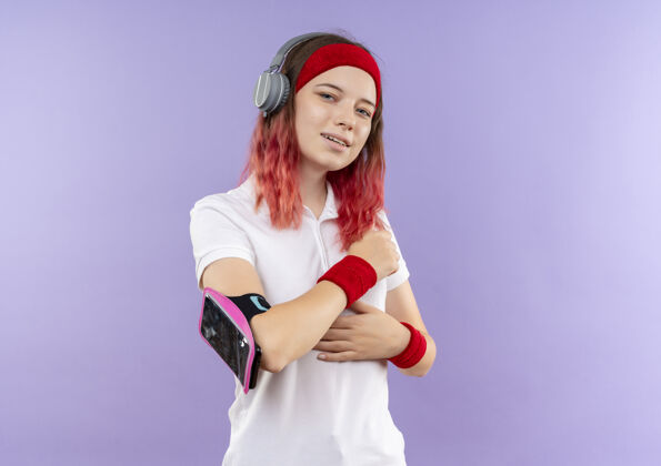 年轻戴着头戴式耳机 戴着智能手机袖标站在紫色墙上微笑的年轻运动女性人运动装人