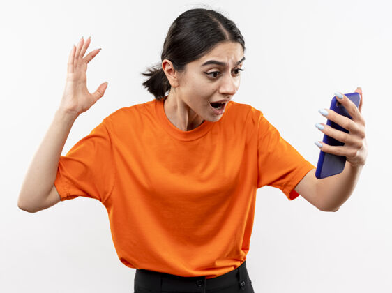 困惑穿着橙色t恤的年轻女孩看着智能手机屏幕兴奋而困惑地举起手站在白色的墙上屏幕移动手