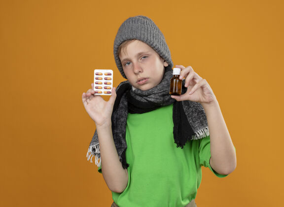 疾病生病的小男孩穿着绿色t恤 戴着暖和的围巾和帽子 感觉不舒服 站在橙色的墙上 展示着药瓶和药丸 不开心 生病围巾看小