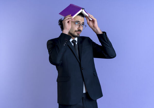 男人一个穿着黑西装戴着眼镜的商人站在蓝色的墙上 头上举着笔记本 看上去又累又无聊眼镜抱商务