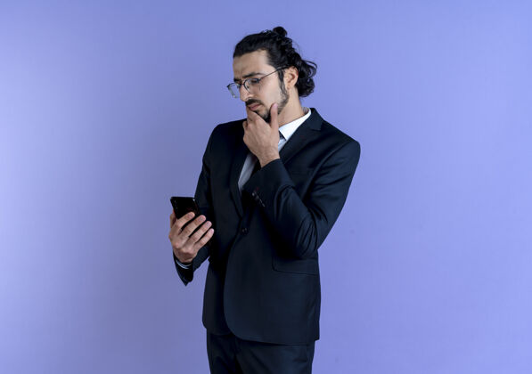 手机一个穿着黑西装戴着眼镜的商人站在蓝色的墙上 脸上带着沉思的表情看着手机屏幕商人市民男人