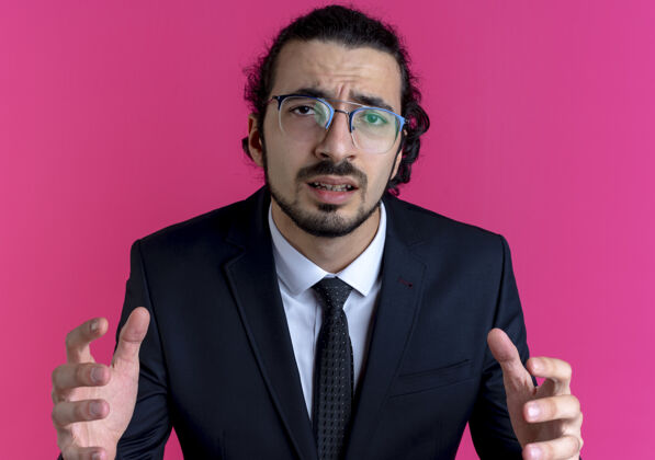 人一个穿着黑色西装戴着眼镜的商人站在粉红色的墙上 困惑地看着前面 举起双臂姿势男人生意人