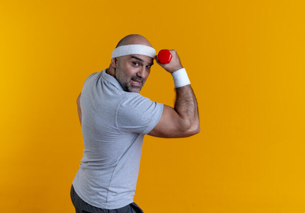 肌肉戴着头巾的成熟运动型男子站在橙色的墙上举手 哑铃看起来紧张而自信提高男看