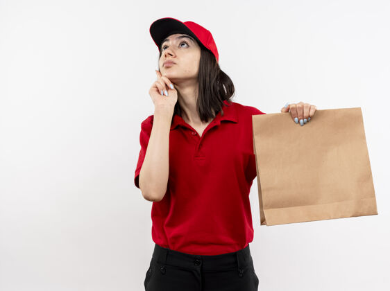抱年轻的送货员身穿红色制服 戴着帽子 手里拿着纸包裹 站在白色的墙上 疑惑地抬头望着拼图女孩包装