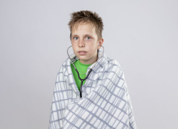 毯子生病的小男孩穿着绿色t恤 裹着毛毯 站在白色的墙上 用听诊器看不舒服站感觉不舒服