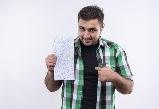 站着穿着格子衬衫的年轻旅行家拿着地图 手指指着地图 站在白色的墙上 看上去很自信年轻人公民地图