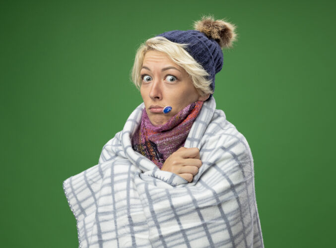 短裤生病的不健康的妇女 短头发 戴着暖和的围巾 戴着帽子 感觉不舒服 裹着毯子 嘴里叼着温度计 站在绿色的背景上温暖包裹不健康
