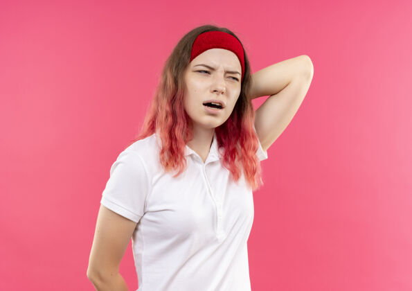 困惑戴着头巾的年轻运动型女人站在粉红色的墙上 困惑地看着一边 摸着脖子 疼得要命站运动运动员