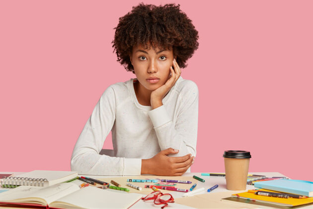 工作一张迷人的黑人设计师的照片 留着非洲发型 坐在工作场所 穿着白色套头衫 用蜡笔画画 隔着粉色的墙壁 享受着咖啡学习室内个人
