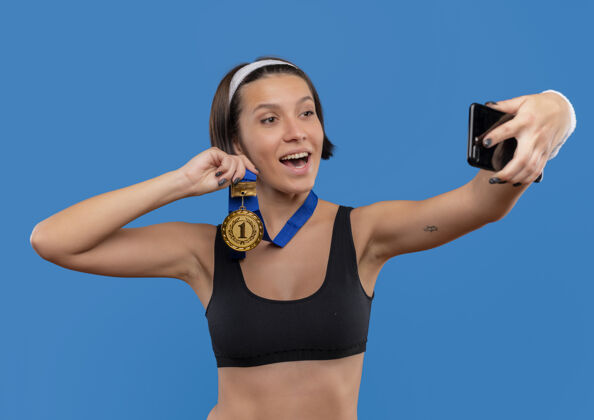 运动身穿运动服 脖子上挂着金牌的年轻健身女士自拍 将金牌展示给站在蓝色墙上的智能手机摄像头自拍人物健康