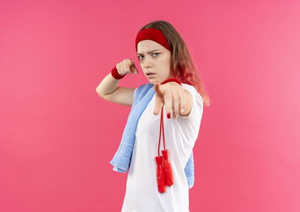 运动装戴着头巾肩上拿着毛巾的年轻运动型女人 手指着相机 严肃的脸站在粉色的墙上人肩膀指尖