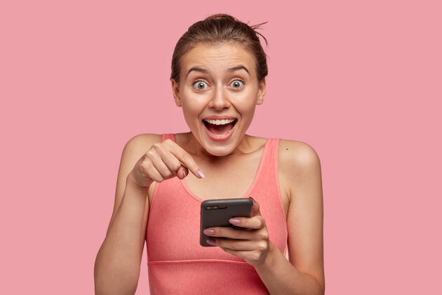 衣服情绪化的白人运动女性照片有着惊讶的表情 用食指指指在手机显示器上 穿着休闲背心 收到良好的通知 隔着粉色的墙壁指向运动成人