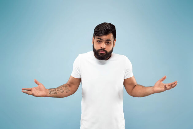工作蓝色背景上穿着白衬衫的印度年轻人的半身特写肖像人类情感 面部表情 广告概念负面空间混乱 未知 不确定专业男人人