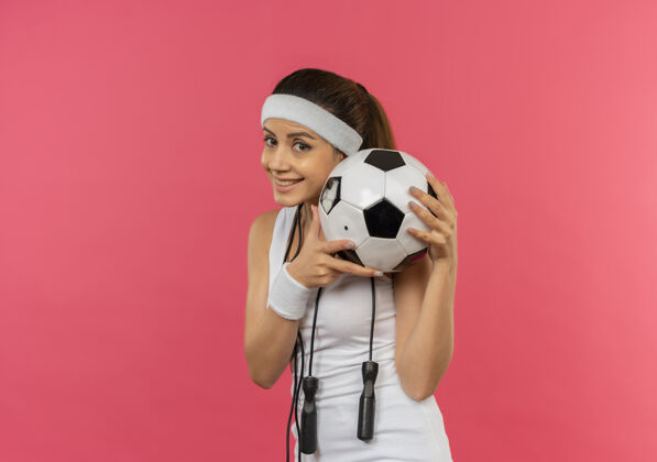 健身身穿运动服 头戴头带 脖子上系着跳绳的年轻健身女士 面带微笑地举着足球站在粉色的墙上脸周围姿势