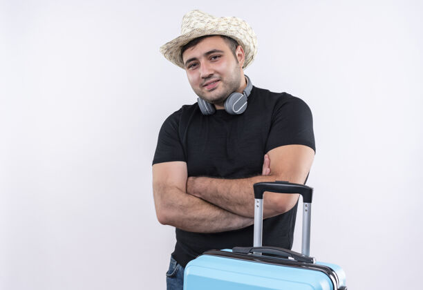 人一个年轻的旅行者 穿着黑色t恤 戴着夏天的帽子 挎着手提箱 双臂交叉 自信地站在白色的墙上年轻人手臂站