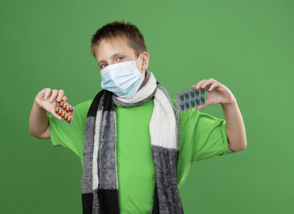周围生病的小男孩穿着绿色t恤 围着暖和的围巾 戴着护面面具 看着站在绿色背景上的摄像机保护疾病面具