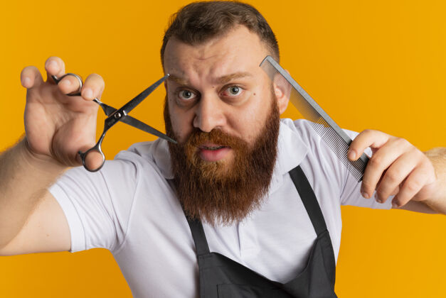 人围裙上的专业胡须理发师拿着剪刀和发刷站在橙色的墙上剪姿势剪站