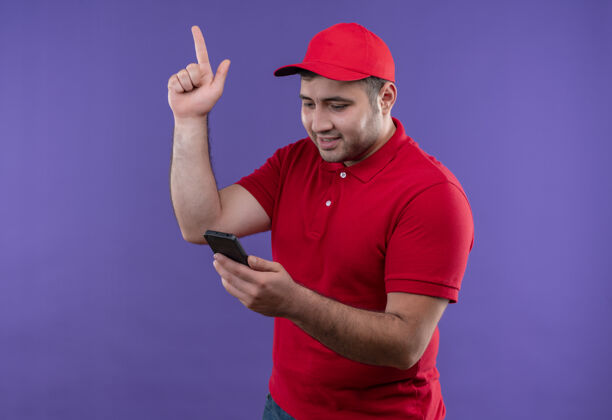 年轻身穿红色制服 戴着帽子的年轻送货员站在紫色的墙上 看着智能手机屏幕 指着有着新的伟大想法的手指公民站看