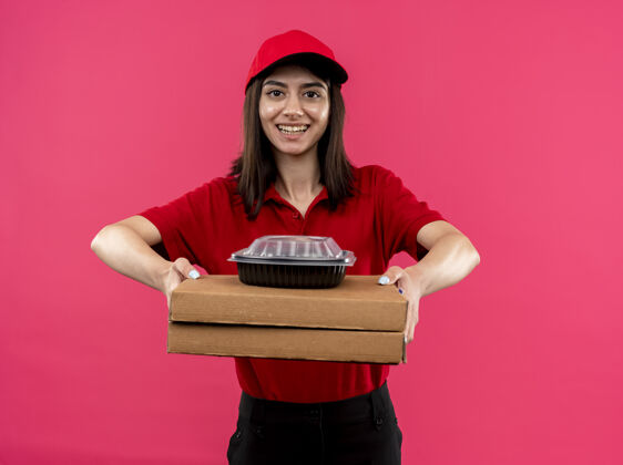 马球身穿红色马球衫 头戴帽子的年轻送货女孩拿着披萨盒和食品包看着镜头 站在粉色背景下 脸上洋溢着幸福的笑容女孩帽子送货