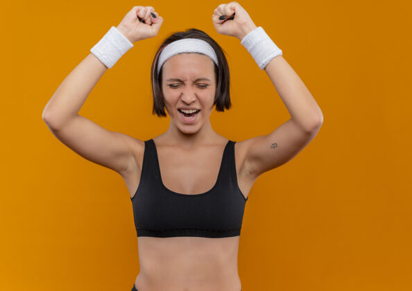 成功身穿运动服的年轻健身女士 头箍紧握拳头 为自己的成功而欢欣鼓舞 疯狂快乐地站在橙色的墙上头带市动