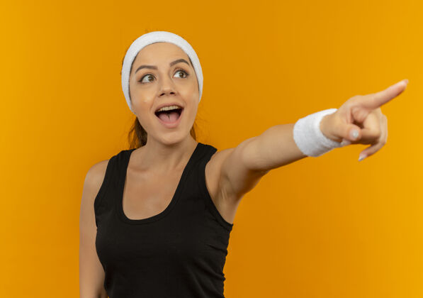 运动员身穿运动服的年轻健身女士戴着头巾 高兴而又惊讶地用手指着站在橙色墙上的一边运动装人人