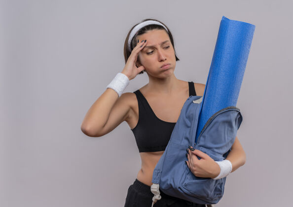 市民年轻的健身女士穿着运动服 背着背包 拿着瑜伽垫 站在白色的墙上 茫然而疲惫地吹着脸颊吹女人健身