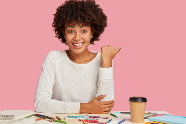 自由职业者快乐积极的黑皮肤女性 非洲发型 为您的广告或促销活动留出自由空间 有愉快的面部表情 喜欢创造性的工作 在粉红色的墙上模特桌面民族人