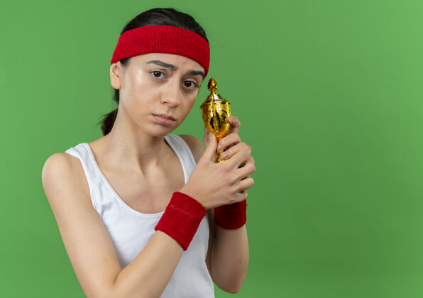 人身穿运动服 头戴钢带 手持奖杯 脸上带着悲伤表情的年轻健身女士站在绿色的墙上运动运动员站着