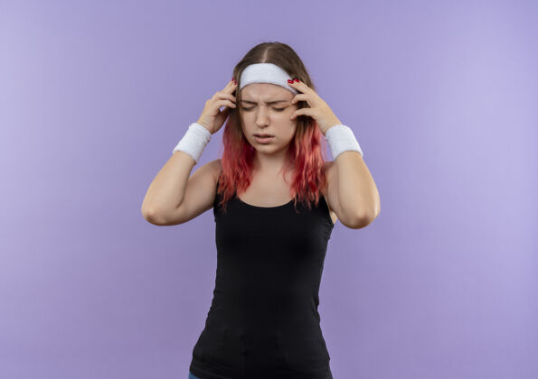 健身穿着运动服的年轻健身女士站在紫色的墙上 看着不舒服 摸着头痛的头女人年轻市民