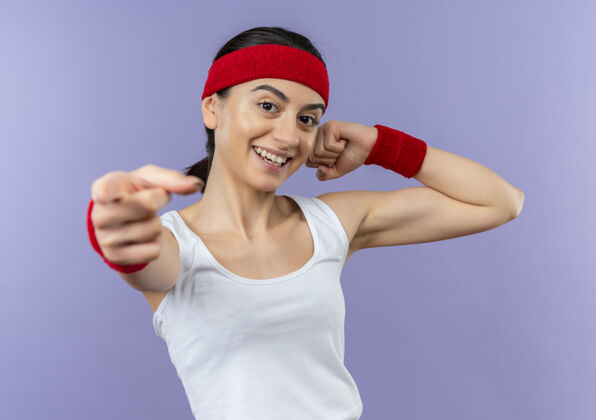 站立身穿运动服的年轻健身女 头戴头巾 举起拳头 手指对着镜头微笑 自信地站在紫色的墙上手指运动头带