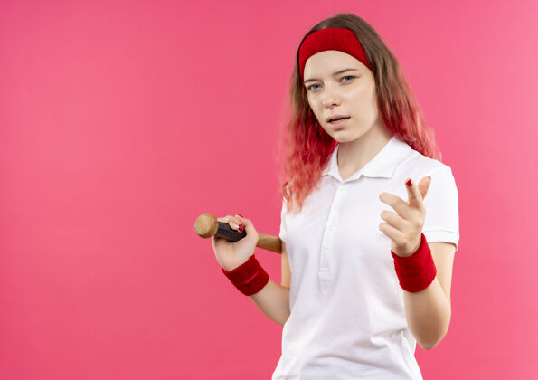 自信戴着头巾 手持棒球棒 自信满满的年轻运动女性站在粉色墙壁上 手指着相机人公民手指