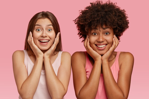 同伴快乐美丽的多民族女孩抱着下巴 接受朋友的好消息 笑容灿烂 在粉红色的墙壁上做模特 表达愉快的情感跨种族关系概念满意牙齿位置