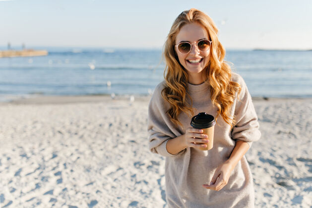 装备迷人的女人 卷发 在沙滩上喝茶时髦的女人 穿着毛衣 在沙滩上放松秋日天空假期金发