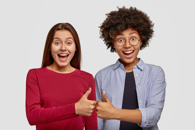 女人两个女人互相竖起大拇指 表示同意 高兴地微笑 肩并肩地站在白墙上 在室内做手势异族朋友高兴 给行手势 喜出望外Excel多样性观点
