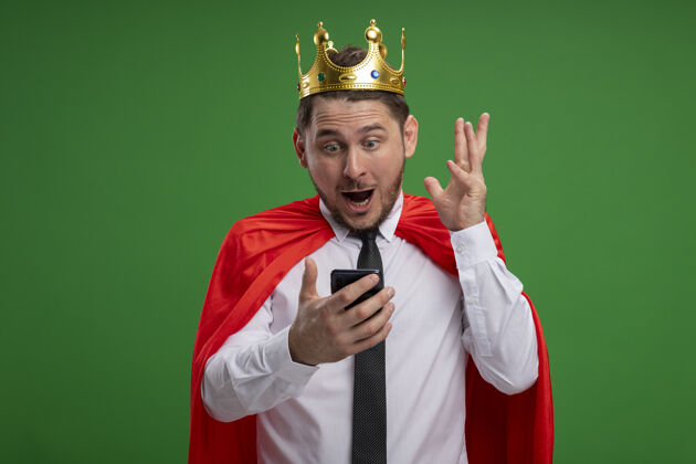 惊人穿着红色斗篷戴着王冠的超级英雄商人站在绿色的背景下 用智能手机看着惊奇和惊讶超级英雄移动惊喜