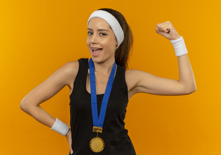 运动身穿运动服 头上戴着头带 脖子上挂着金牌的年轻健身女士高举拳头 站在橘色的墙上 开心而积极地微笑着健身人周围