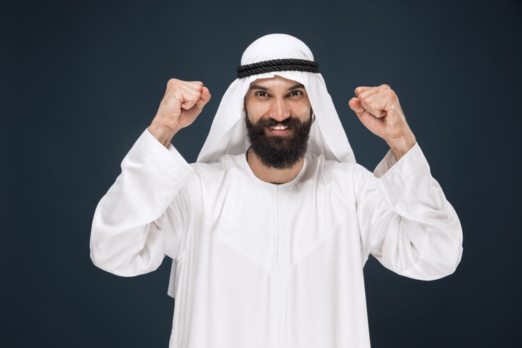情感深蓝色工作室背景上的阿拉伯沙特商人肖像年轻的男模站着 微笑着庆祝商业概念 金融 面部表情 人类情感人类男性发呆
