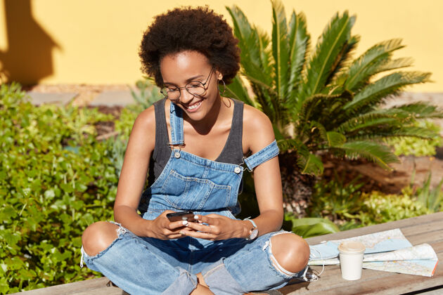 谈话积极的女性游客使用现代手机在社交网络中发送信息 双腿交叉 戴眼镜和牛仔裤 享受热带度假 咖啡和地图在线热带设备