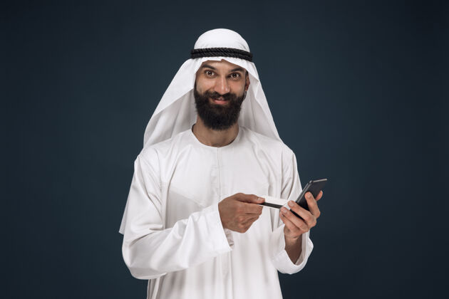 伊斯兰深蓝色工作室背景上的阿拉伯沙特商人肖像男子使用智能手机付账 网上购物或投注商业概念 金融 面部表情 人类情感人类打赌震惊