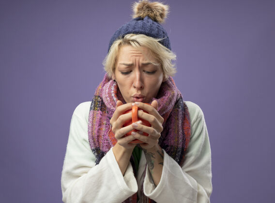 帽子生病的不健康的女人 短头发 戴着暖和的围巾和帽子 感觉不舒服 拿着一杯热茶站在紫色的背景上喝短裤温暖生病