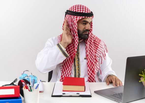 笔记本电脑身着传统服装的阿拉伯商人手拿笔记本电脑工作 耳边试图听到坐在办公桌旁的声音传统试坐