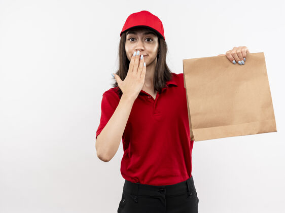帽子年轻的送货女孩穿着红色制服 戴着帽子 拿着纸包看着镜头 惊讶地用手站在白色的背景上举行年轻立场