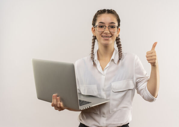 微笑戴着眼镜 梳着辫子 穿着白衬衫的年轻女学生拿着笔记本电脑望着前面 兴高采烈地笑着竖起大拇指站在白墙上看年轻人笔记本电脑
