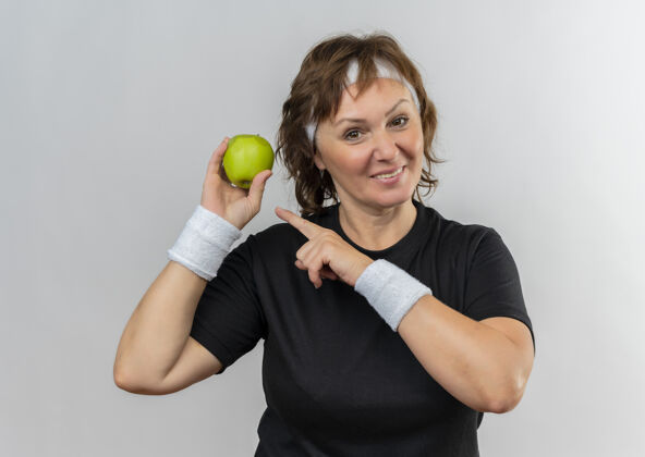 运动装中年运动女性 身穿黑色t恤 头上戴着头巾 手拿两个绿色的苹果 手指着苹果 站在白色的墙上开心地笑着微笑女人人