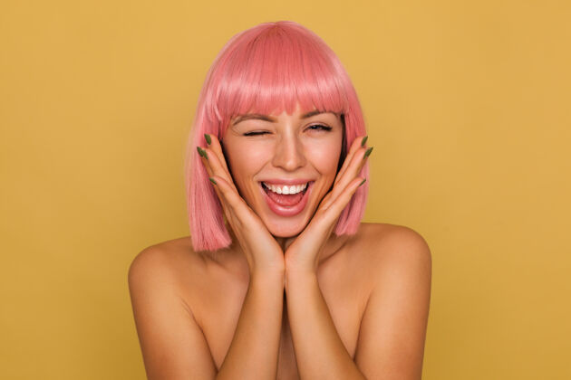 20秒室内拍摄的年轻迷人的粉红色头发的女人与鲍勃的发型使眼色和微笑愉快 而依靠下巴在提高手掌 隔离在芥末墙情感短裤女士