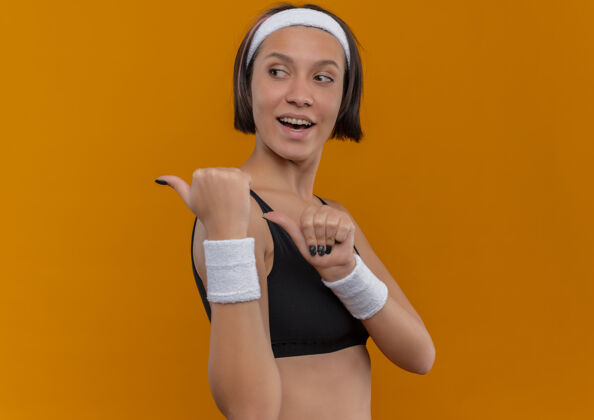 女人穿着运动服的年轻健身女士 头上戴着积极的头带 高兴地向后指着 大拇指站在橙色的墙上背部女人运动员